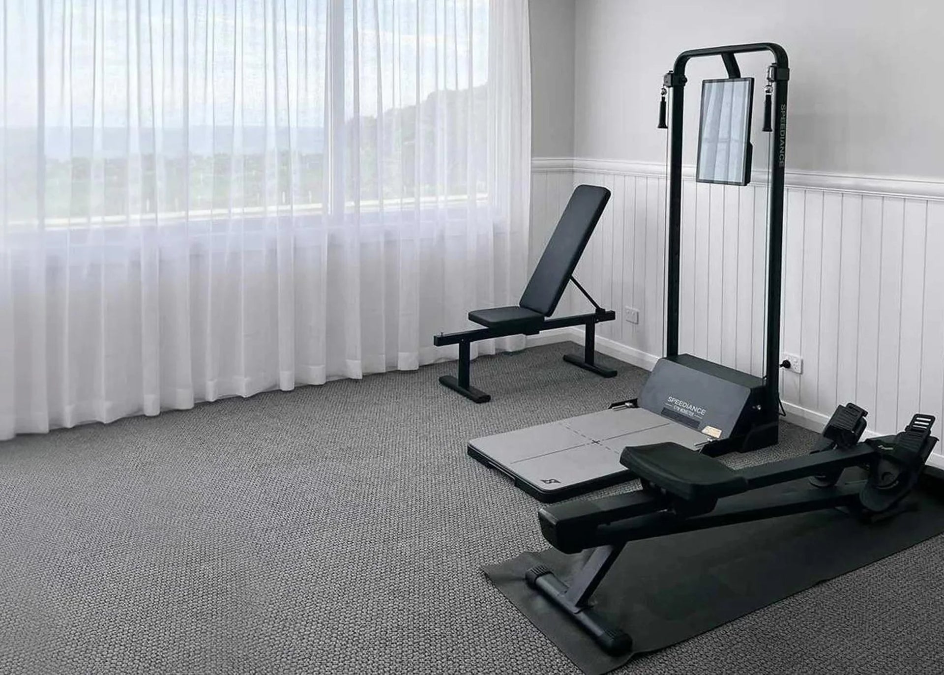 Home gym setup with weight bench, treadmill, and resistance machine in a room with large windows.